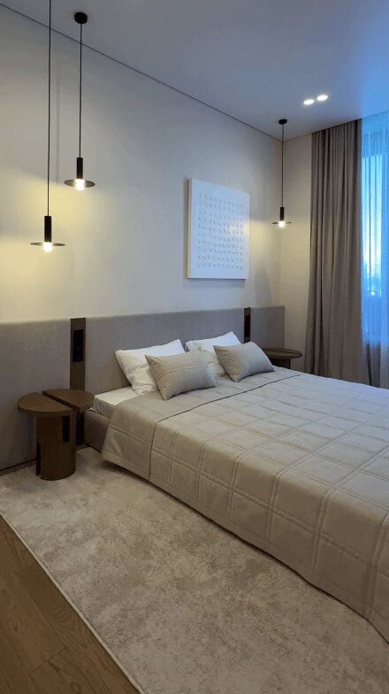 Спальня, ліжко з мяким узголів'ям, круглий приліжкий столик, чорні мінімалістичні світильники підвішані на різній висоті, стіни бежевого кольору