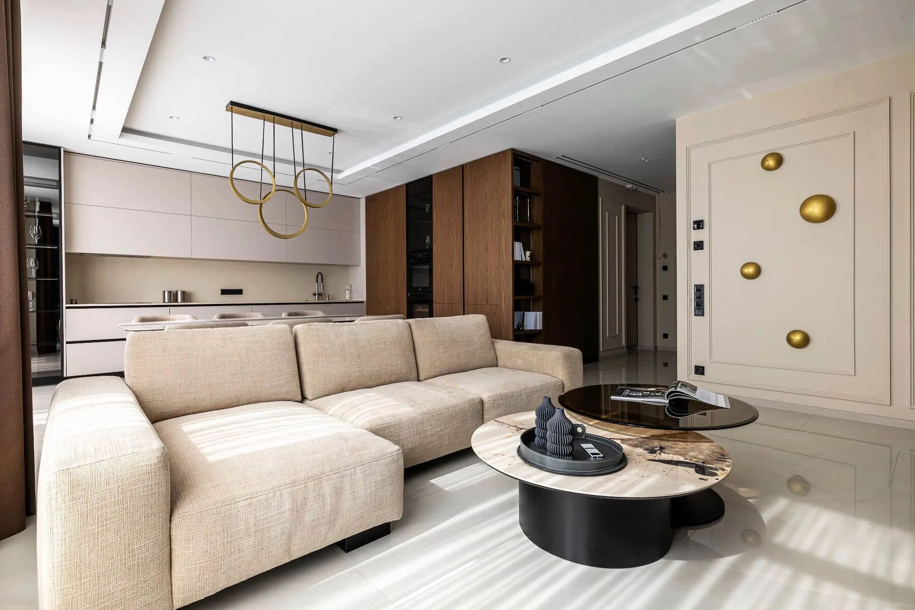 Простора кухня-вітальня, з бежевим диваном, італійським дворівневим журнальним столиком, дизайнерським світильником у формі трьох кілець