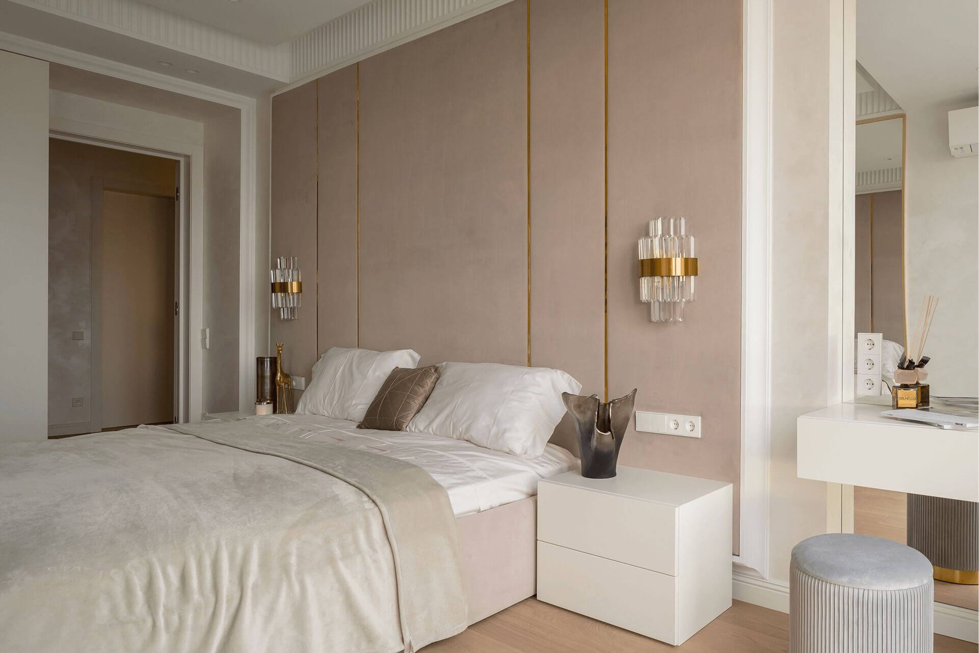 Інтер'єр спальні в класичному стилі з м'якими стіновими панелями персикового кольору за узголів'ям, білими мінімалістичними приліжковими тумбами, настінними бра і туалетним навісним столиком біля дзеркала і сірим пуфом