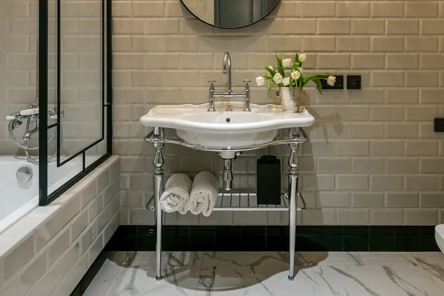 Ванная кімната з білим умивальником на витончених металевих ніжках, білою плиткою та білими трояндами у сріблястій підставці